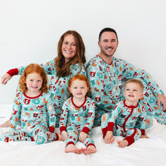 A family of five sits on a bed, all wearing matching holiday-themed pajamas. The group consists of two adults and three young children with various expressions of joy and contentment on their faces.