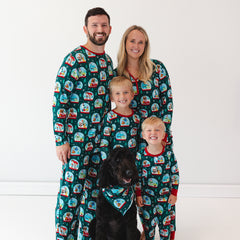 A family of four and a black dog are dressed in matching holiday-themed pajamas featuring colorful designs. The two children are standing in front of the parents, and the dog wears a matching bandana.