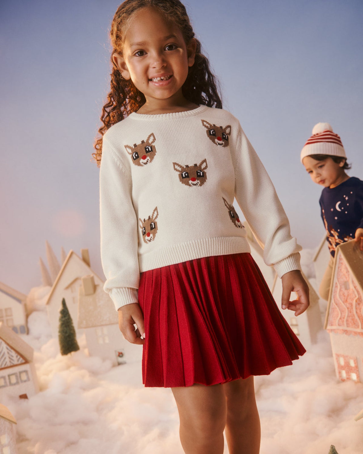 A young girl stands in a snowy village display wearing a cream knit sweater with embroidered reindeer faces and a red pleated skirt. Another child is visible in the background near gingerbread houses under a soft blue sky.