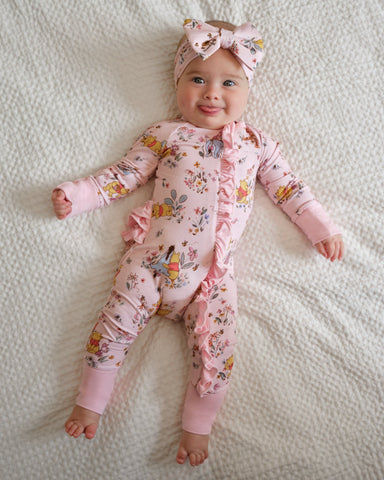 A baby lying on a white textured blanket, wearing a pink long-sleeve footless onesie with a floral and fairy print, ruffle trim, and a matching pink bow headband.