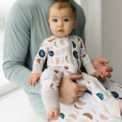 Image of an adult male in solid long sleeve pajama top in Stormy color paired with Luna Neutral printed pajamas. Man is holding an infant in Luna Neutral long sleeve zipper pajamas.