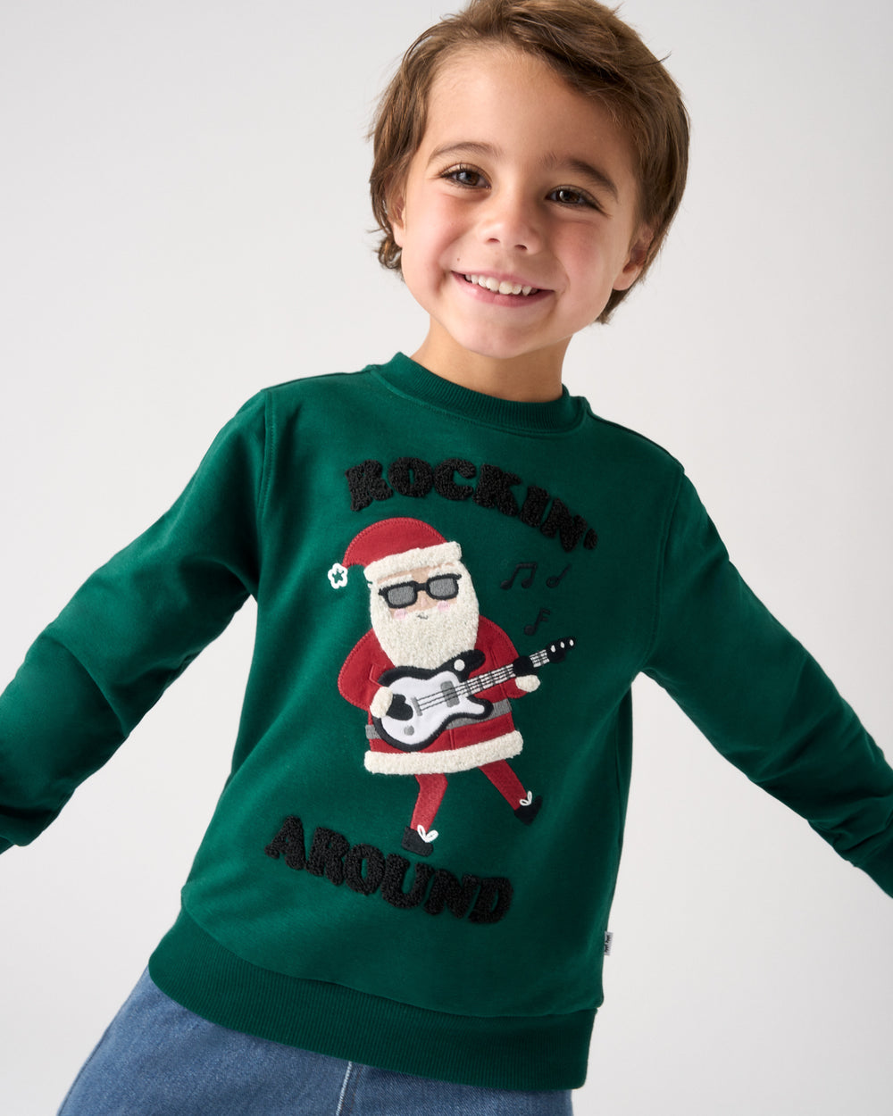 A smiling young boy wearing a dark green crewneck sweatshirt with a graphic of a Santa Claus playing a guitar and the words 'ROCKIN' SANTA AROUND' in black.