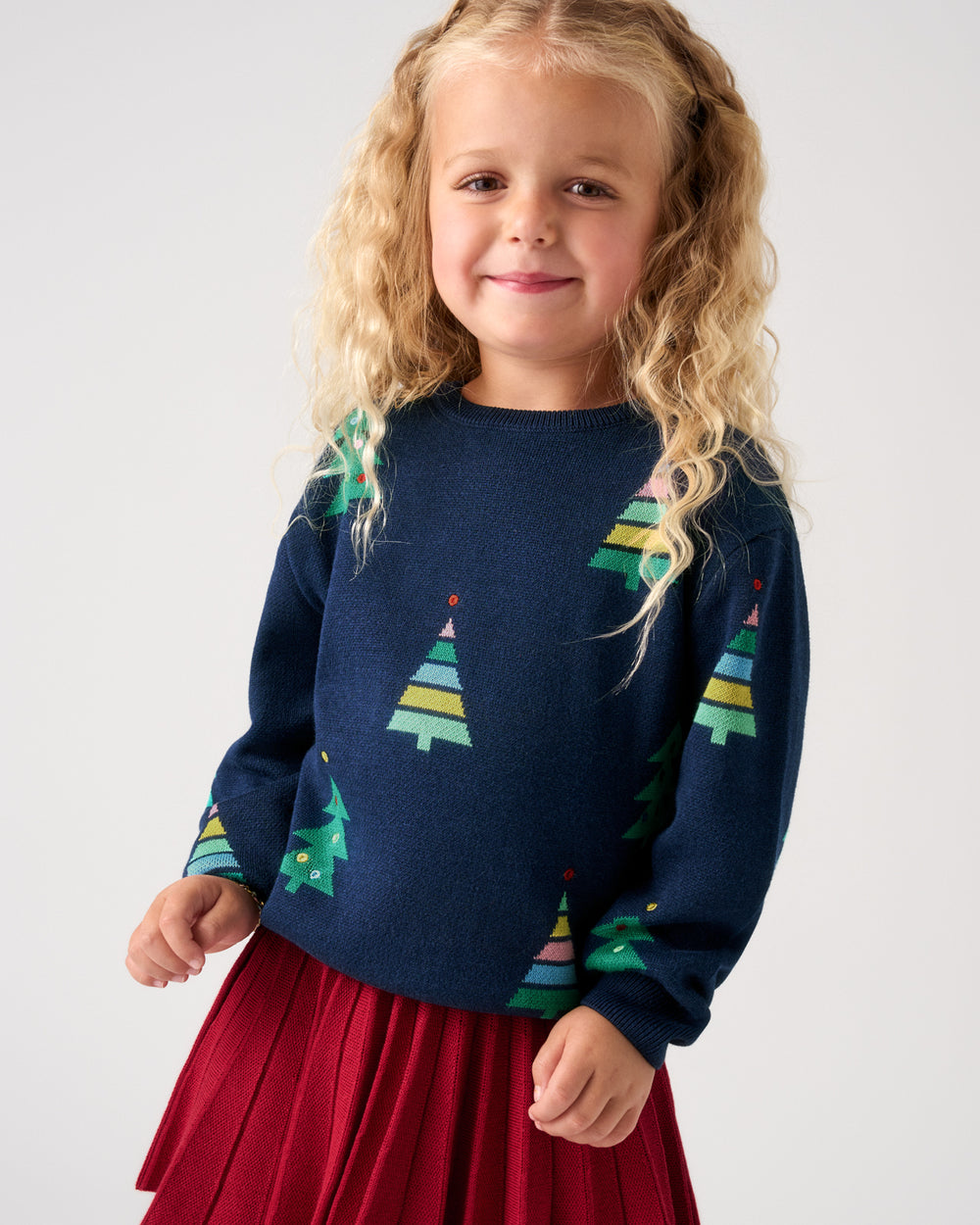 A young girl with curly blonde hair wearing a navy blue knit sweater with a colorful Christmas tree pattern and a red pleated skirt, photographed against a light gray studio background.
