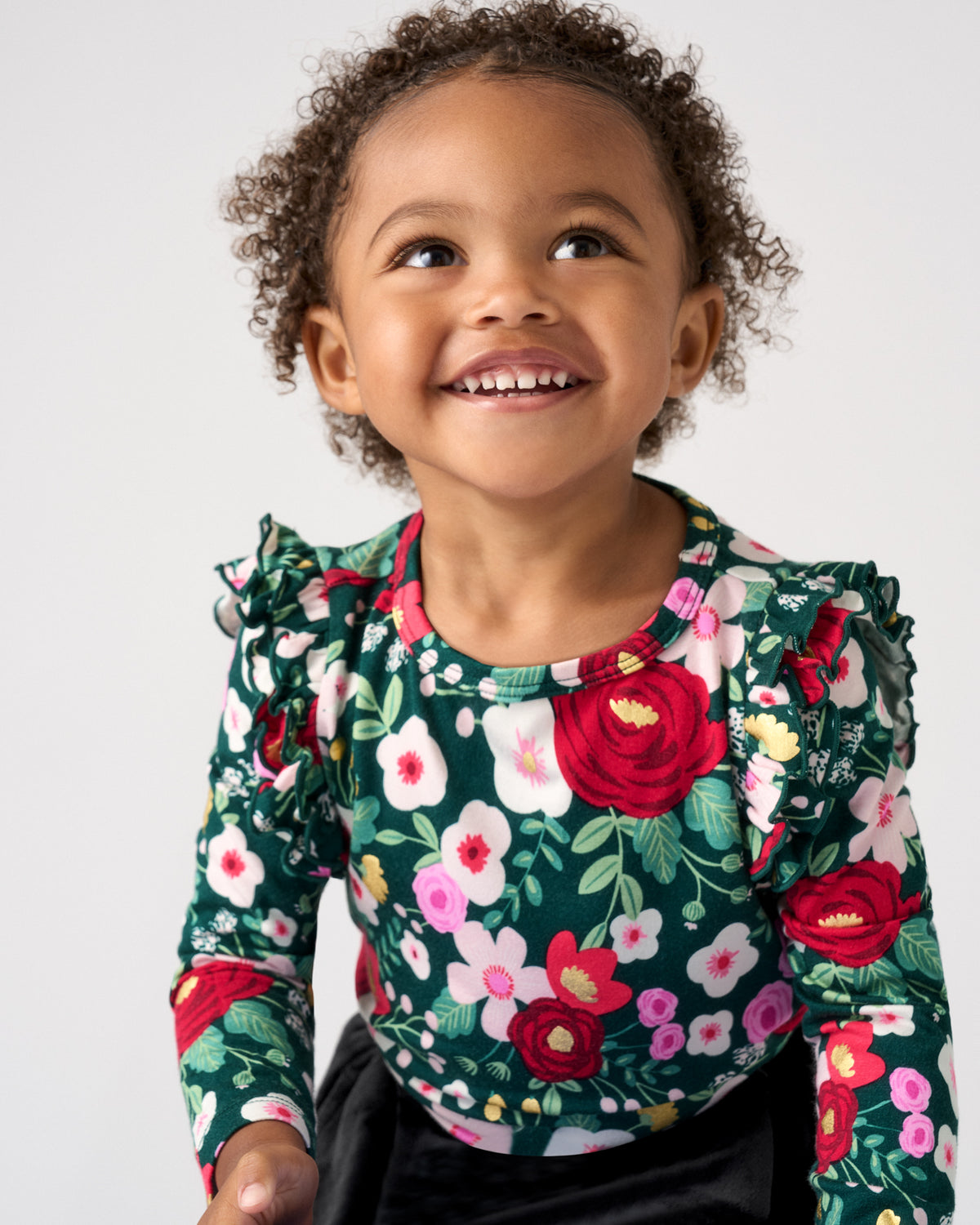 A smiling toddler with curly brown hair wearing a dark green long-sleeve top with a vibrant red, pink, and white floral print, photographed against a solid light gray background.