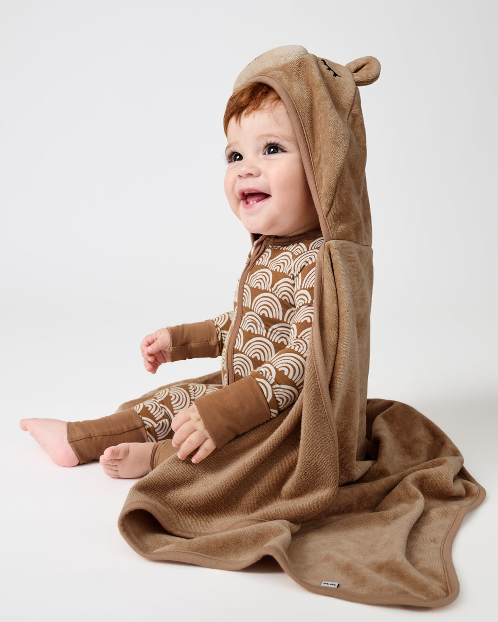 A baby wearing a brown hooded one-piece outfit with a beige and brown wave pattern, sitting on a white studio background. The outfit includes a plush hood with bear ears and a matching brown blanket draped over the baby.
