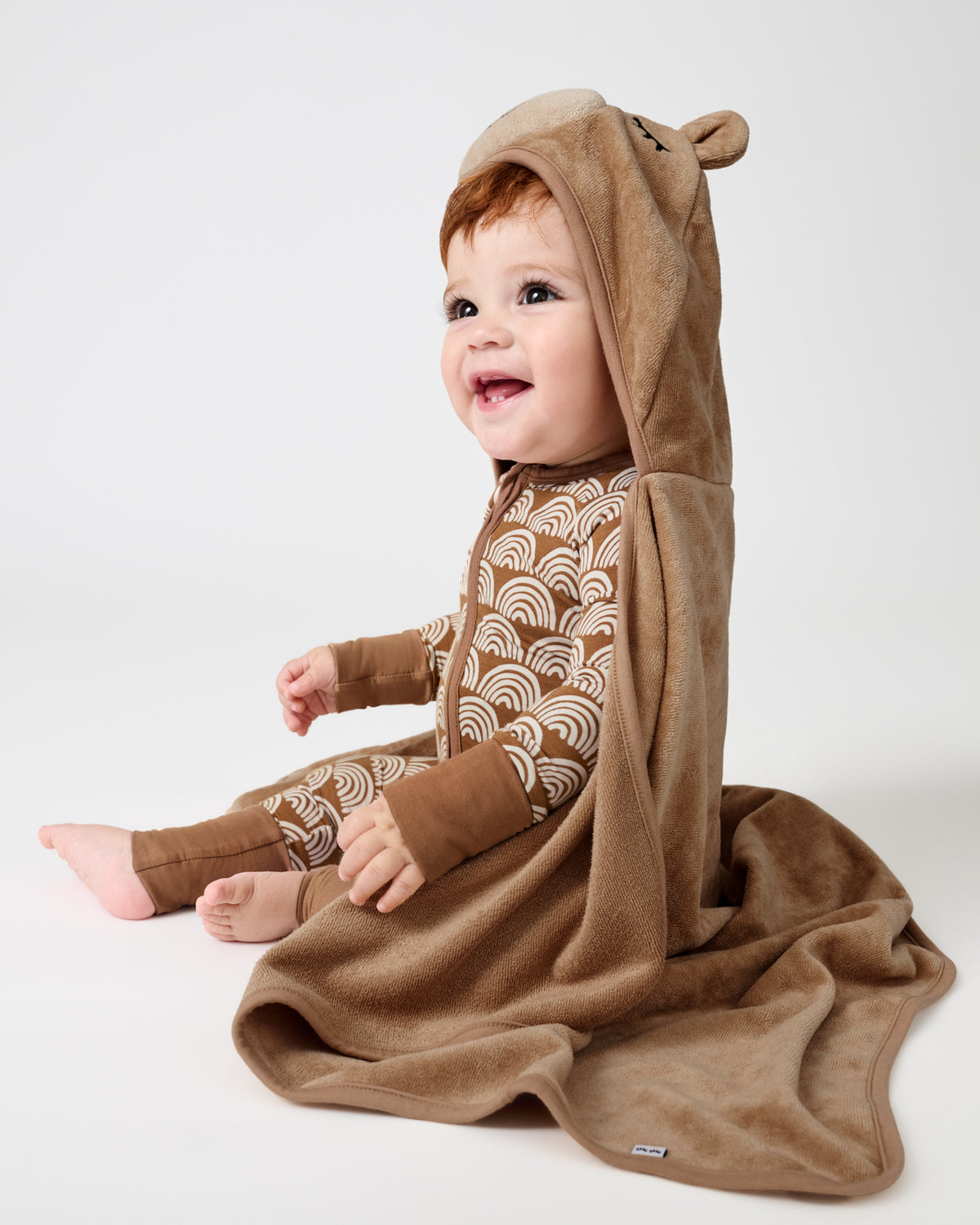 A baby wearing a brown hooded one-piece outfit with a beige and brown wave pattern, sitting on a white studio background. The outfit includes a plush hood with bear ears and a matching brown blanket draped over the baby.