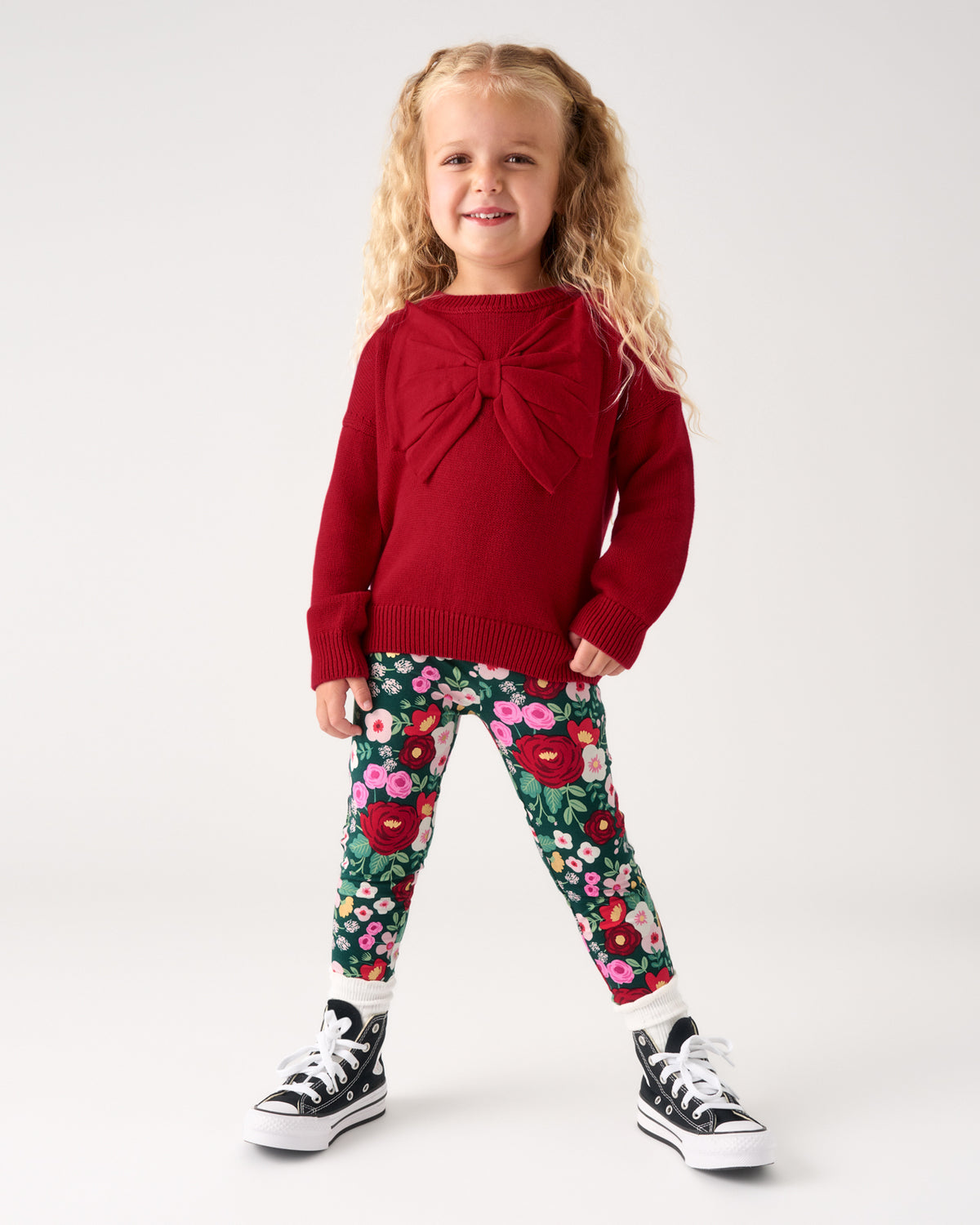 A young girl with blonde curly hair stands against a light gray studio background wearing a dark red knit sweater with a large front bow, floral print leggings, and black sneakers.