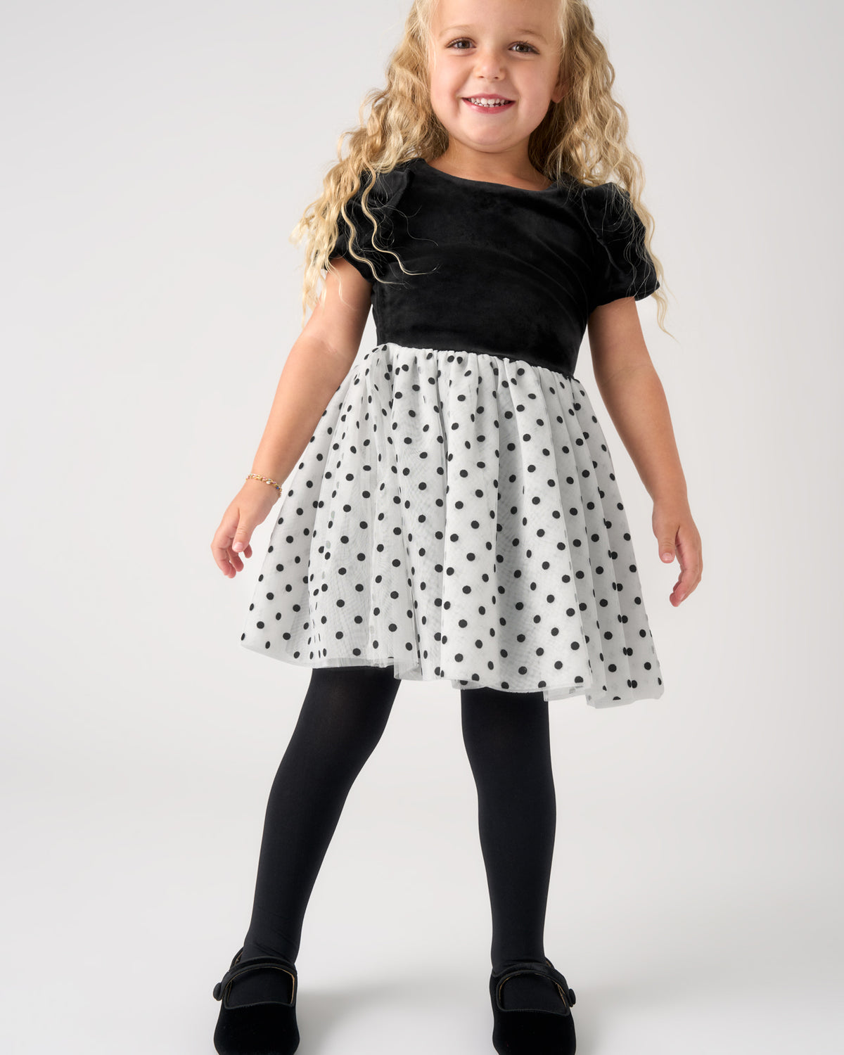 A young girl with long blonde curly hair is modeling a short-sleeve dress. The dress features a black velvet-like bodice and a white flared skirt with black polka dots.