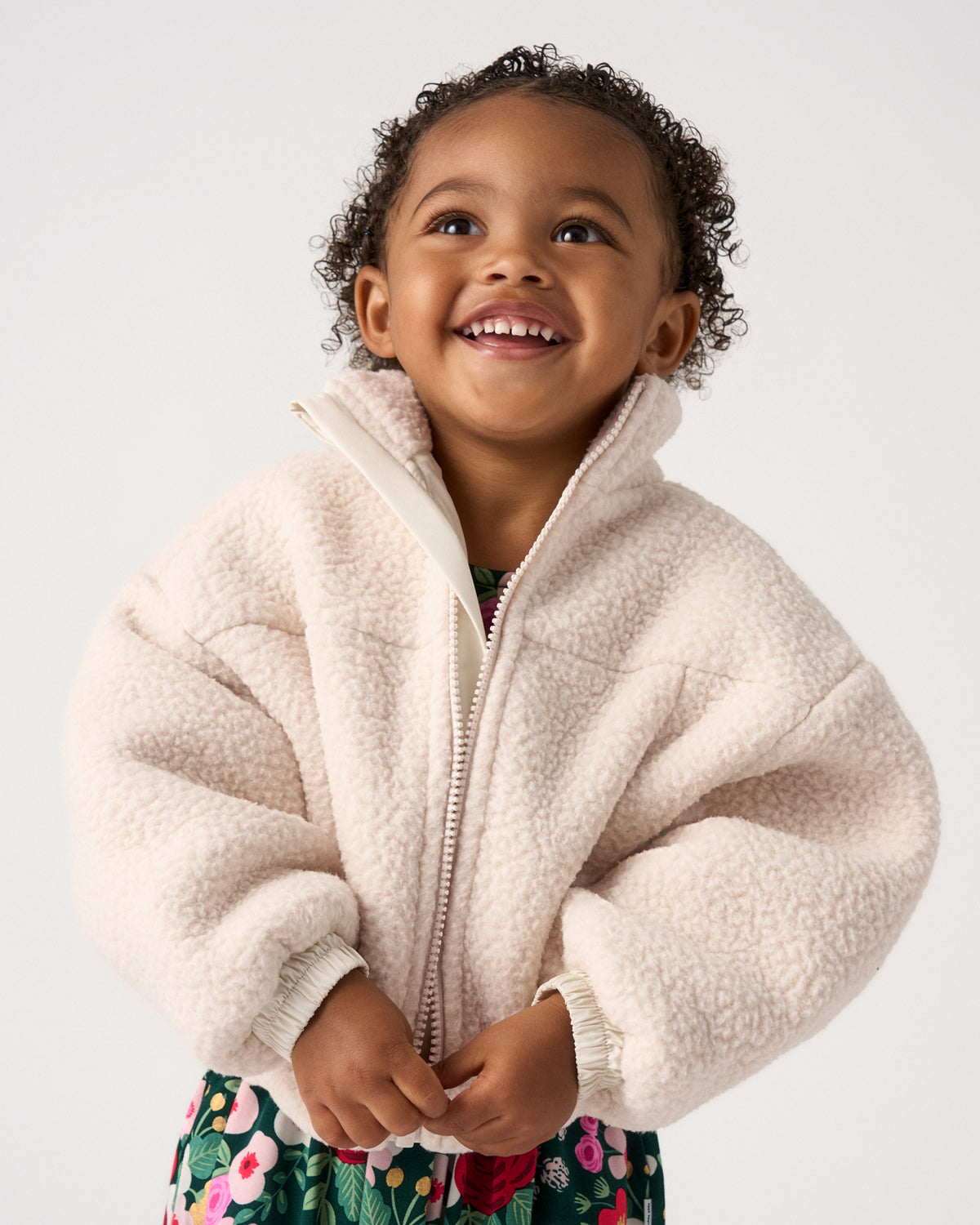 A smiling young child wearing a cream sherpa fleece zip-up jacket with ribbed cuffs and collar, paired with a colorful floral skirt, photographed against a clean light gray studio background.