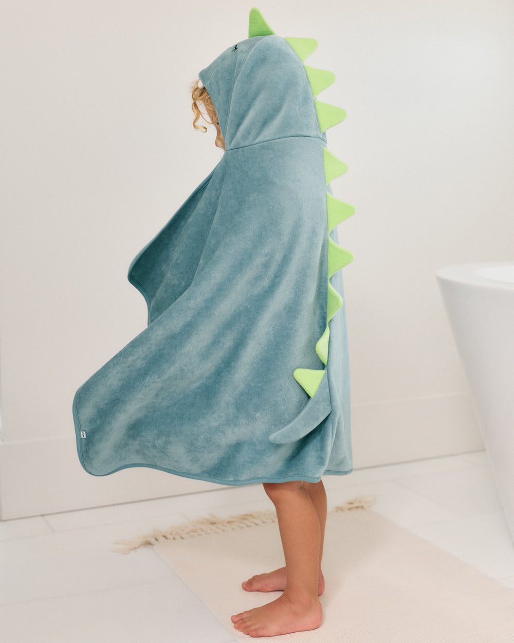 A child wearing a plush dinosaur-themed hooded towel or cape in dusty blue, standing barefoot in a bright bathroom. The hood has a green dinosaur head detail, and the back features a row of green spikes.