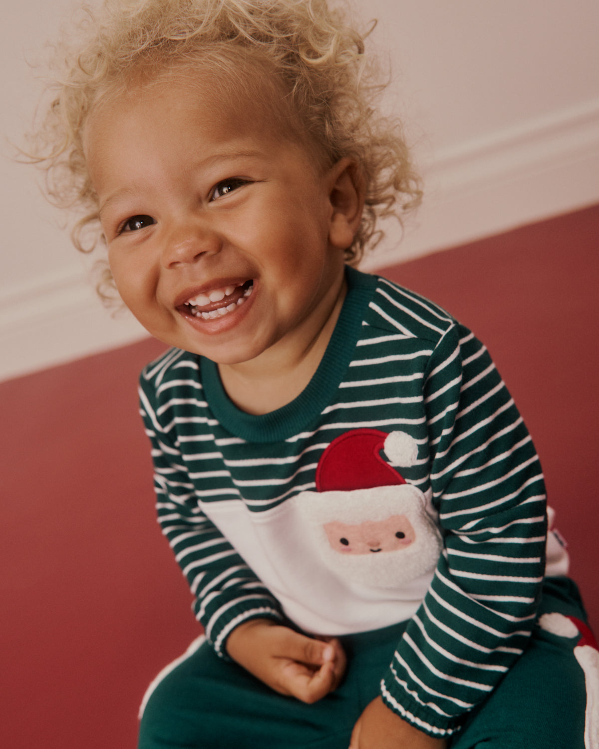 A smiling toddler with curly blonde hair wearing a festive holiday pajama set. The long-sleeve top features dark green and white horizontal stripes, a white chest panel with a Santa Claus face, and a red Santa hat applique.