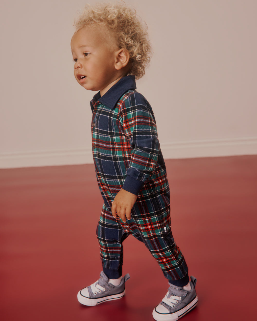 A toddler with curly blonde hair wearing a navy blue plaid jumpsuit with red, green, and yellow lines, walking on a red floor against a beige wall.