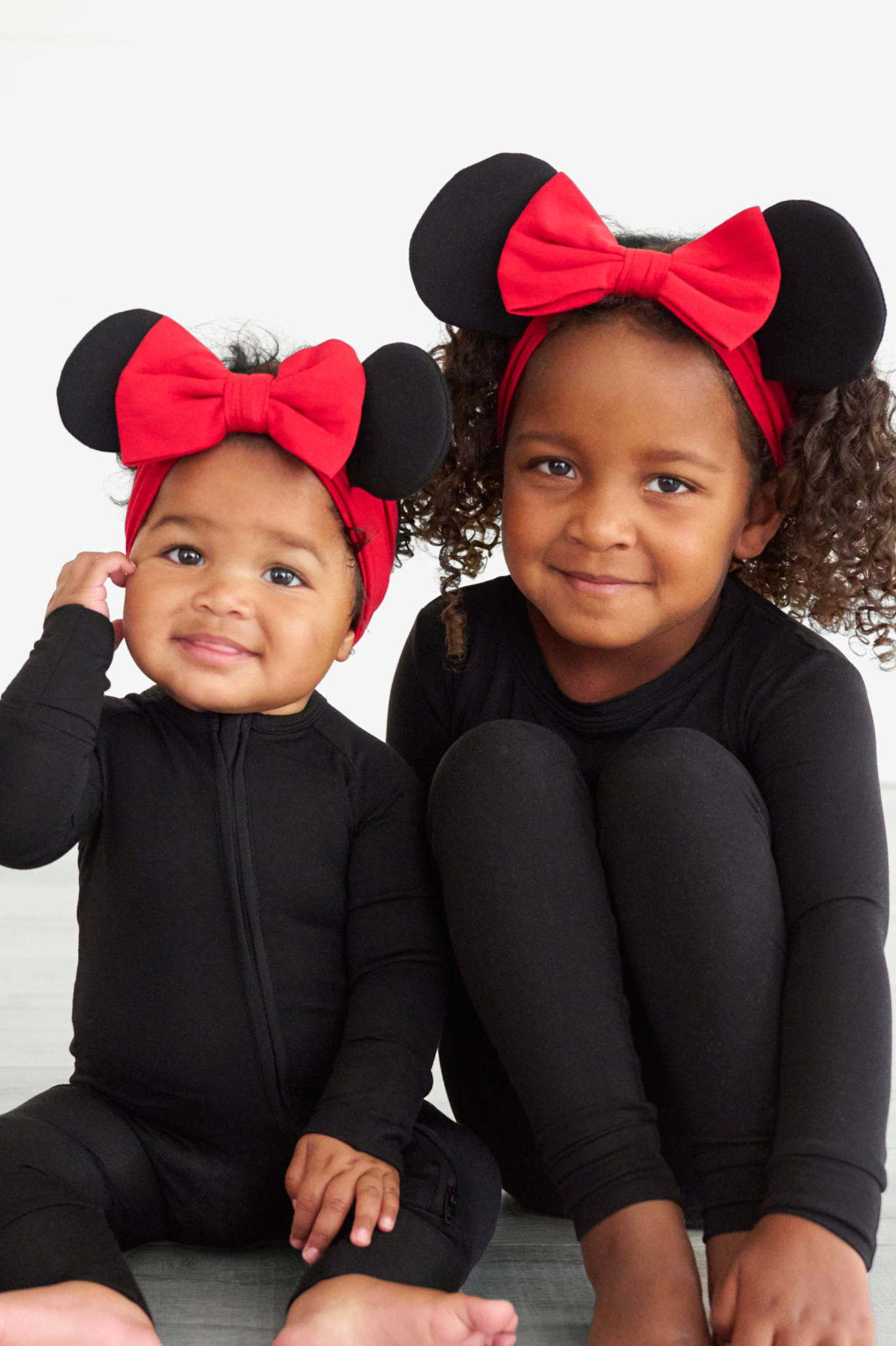 two children sitting on the ground wearing matching black pajamas and red minnie mouse ears luxe bow headbands