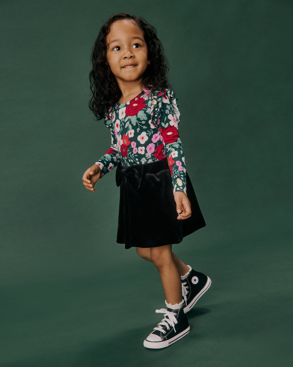 A young girl with curly dark hair stands against a solid dark green studio background, wearing a long-sleeve floral print top with red, pink, and white flowers on a green base, paired with a black velvet skirt and black high-top sneakers.
