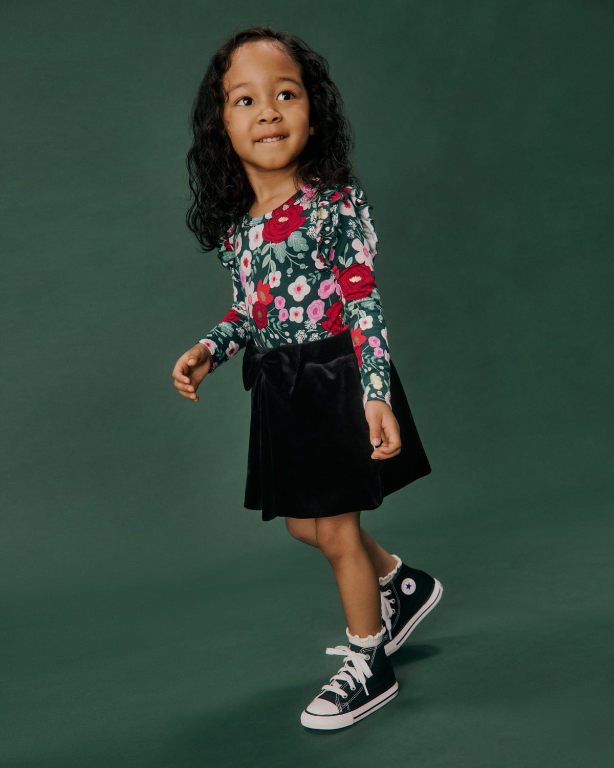 A young girl with curly dark hair stands against a solid dark green studio background, wearing a long-sleeve floral print top with red, pink, and white flowers on a green base, paired with a black velvet skirt and black high-top sneakers.