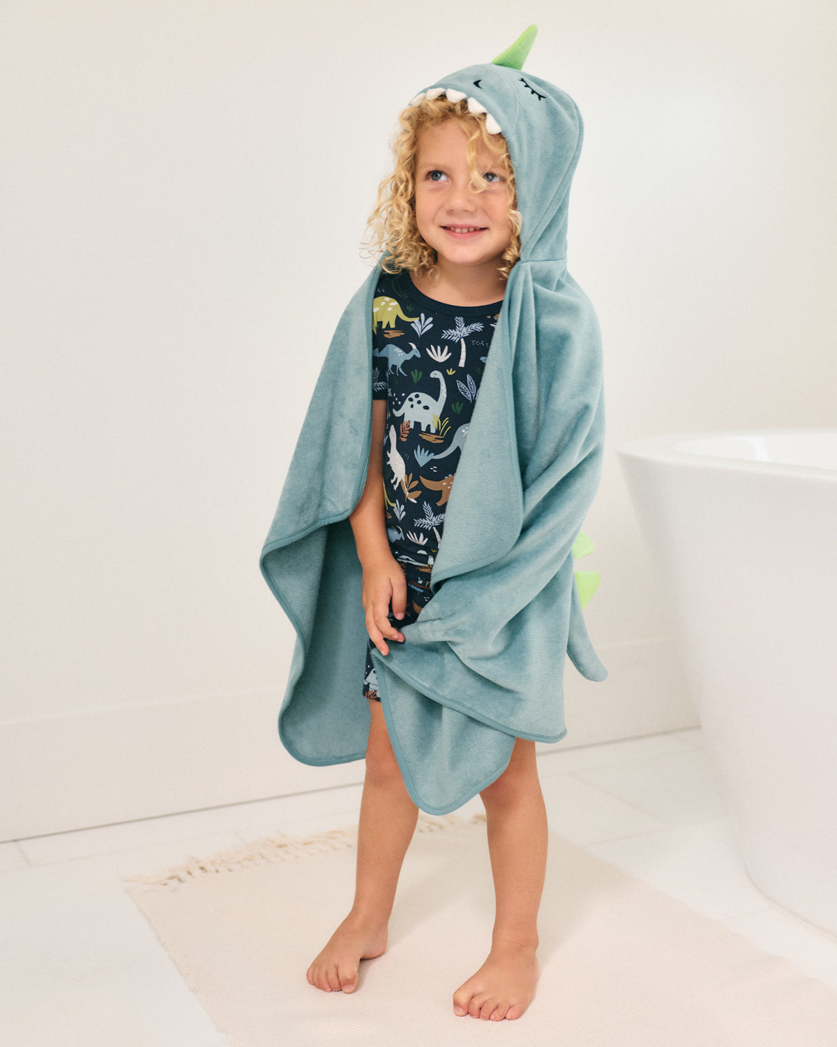 A young child wearing a light blue dinosaur-themed hooded towel wrap, standing barefoot in a bright bathroom next to a white bathtub.