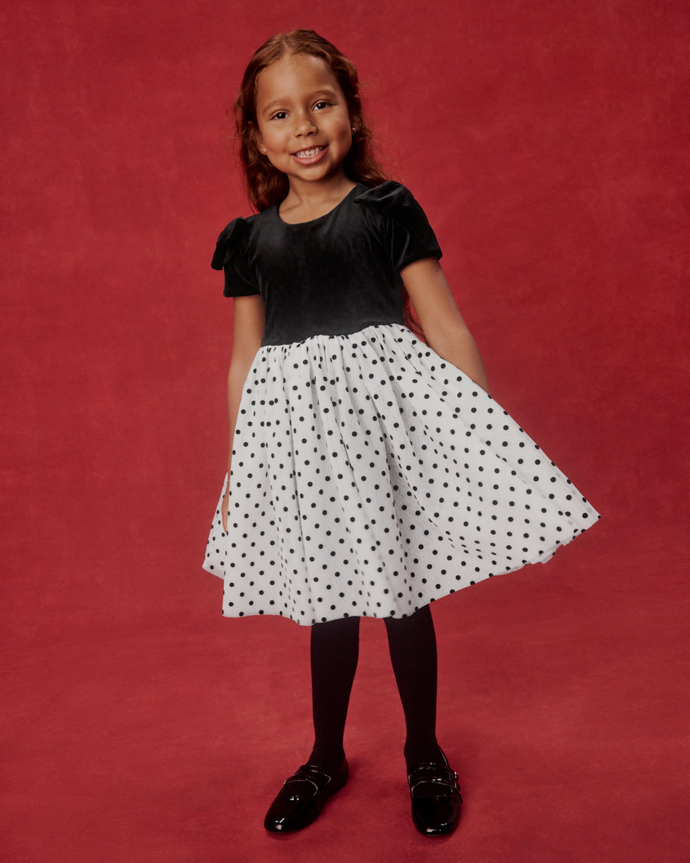 A young girl modeling a black and white polka dot dress against a solid red studio backdrop. The dress features a black velvet short-sleeve bodice and a white A-line skirt with black polka dots.