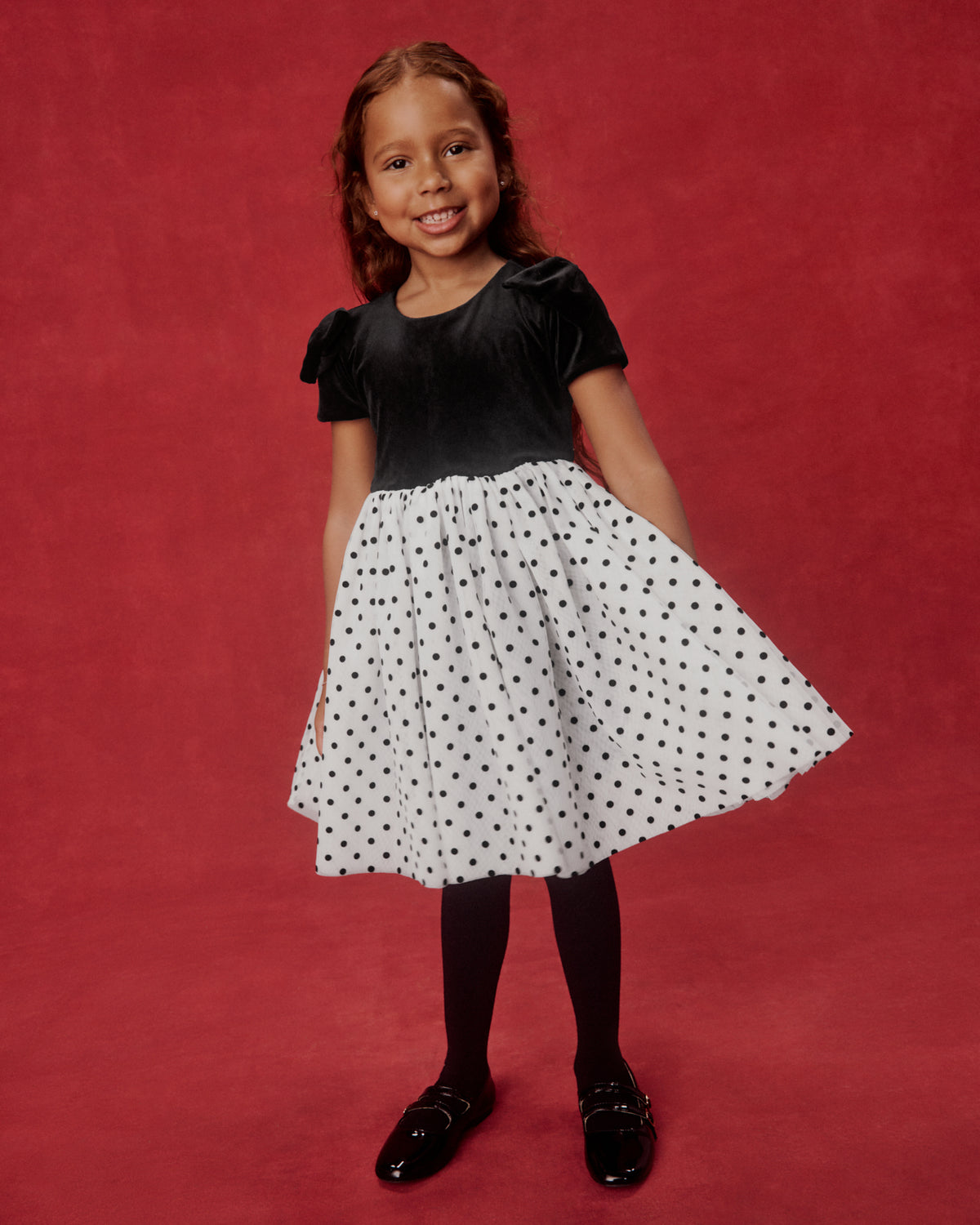 A young girl modeling a black and white polka dot dress against a solid red studio backdrop. The dress features a black velvet short-sleeve bodice and a white A-line skirt with black polka dots.