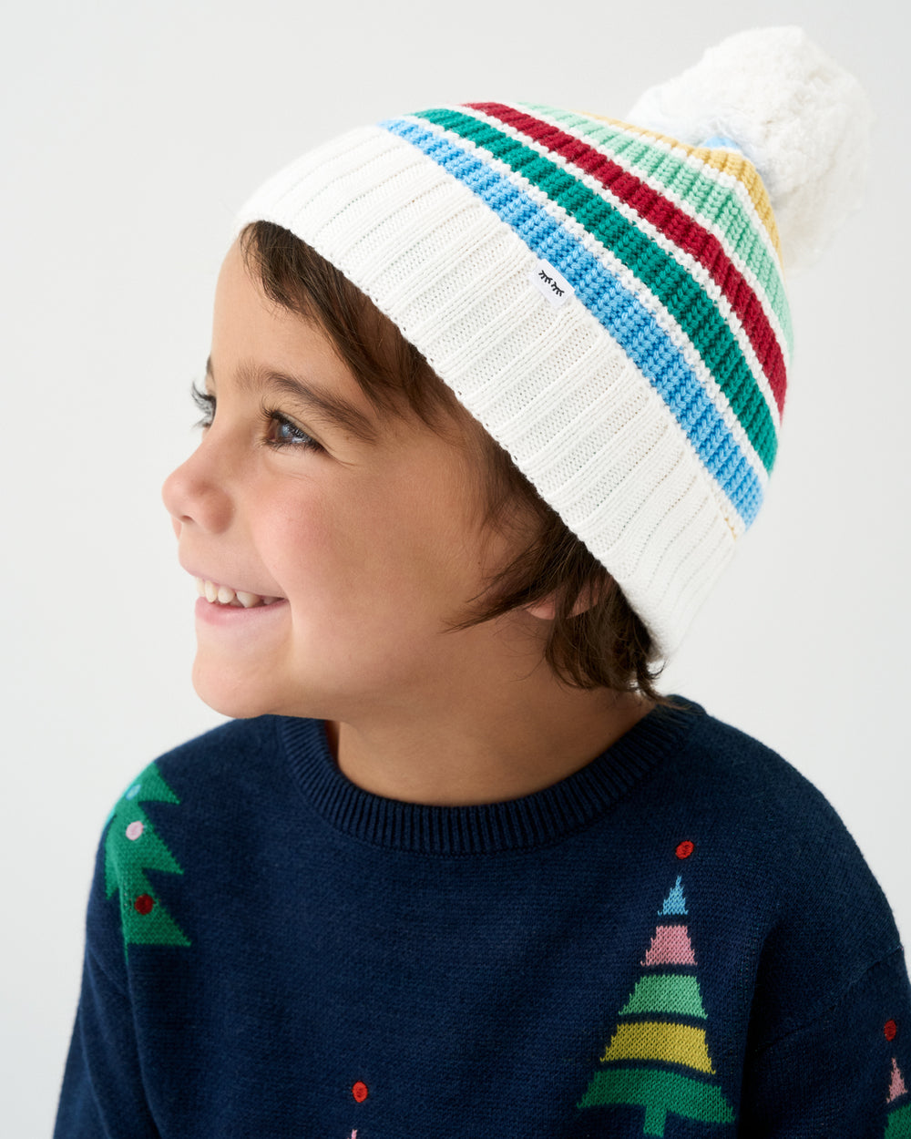 A smiling child in profile wearing a white knit beanie with colorful diagonal stripes and a fluffy pom-pom, paired with a navy sweater featuring Christmas tree patterns.