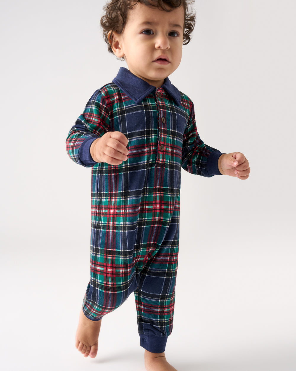 A toddler wearing a long-sleeve plaid romper with a navy collar, standing against a light gray studio background.