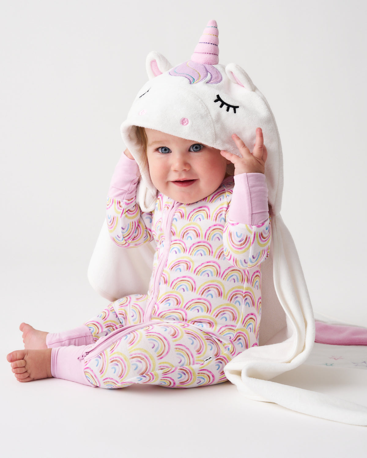 A baby wearing a white hooded onesie with a pink unicorn horn and ears, sitting on a white studio background. The one-piece has a pastel rainbow scallop pattern and pink cuffs and ankles.