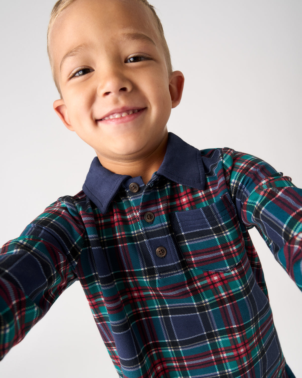 A smiling young boy wearing a long-sleeve plaid button-up shirt with a navy collar, photographed against a plain light gray background.