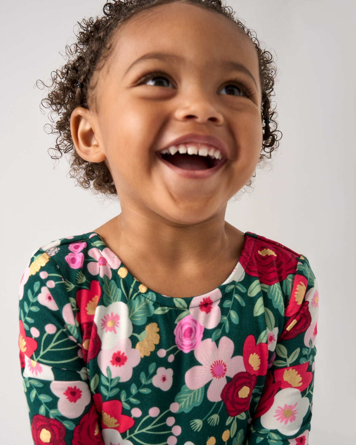 A close-up of a young child with curly brown hair smiling and looking upward, wearing a dark green long-sleeve top with a colorful floral print in pink, red, white, and yellow against a plain light gray background.