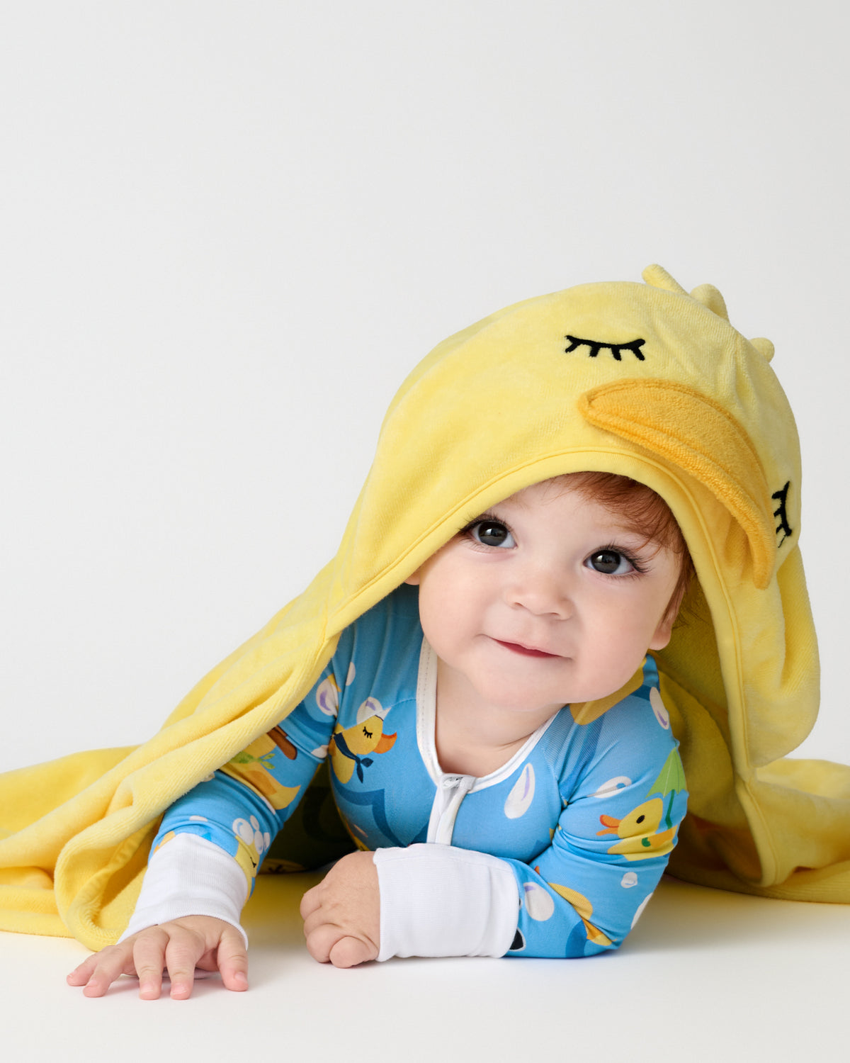 A baby wearing a blue duck-themed long-sleeve onesie and a yellow hooded towel blanket with a duck face, lying on a white surface against a light gray background.