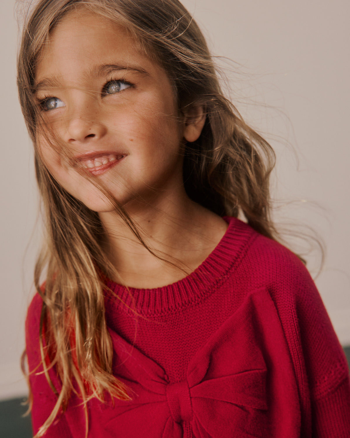 A young girl with long light brown hair looking up and smiling, wearing a bright red knit sweater with a large bow detail at the front.