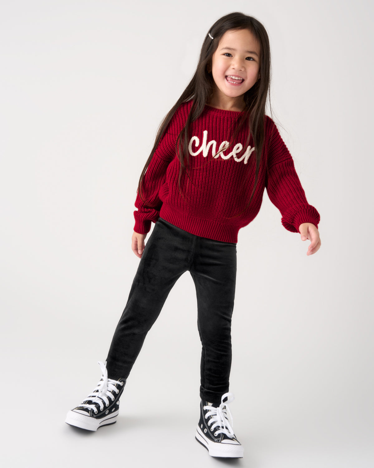 A young girl smiling and posing in a studio, wearing a red ribbed knit sweater with 'cheer' written in white script across the chest, black pants, and black-and-white sneakers.