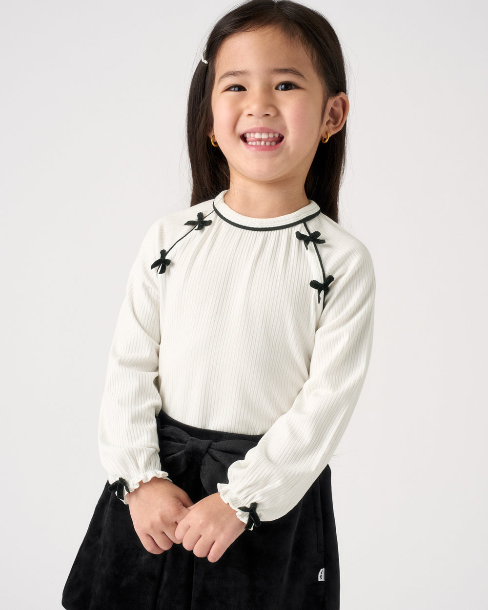 A young girl smiling, wearing a cream ribbed long-sleeve top with black bow details on the shoulders and cuffs, paired with a black skirt, against a plain light gray background.