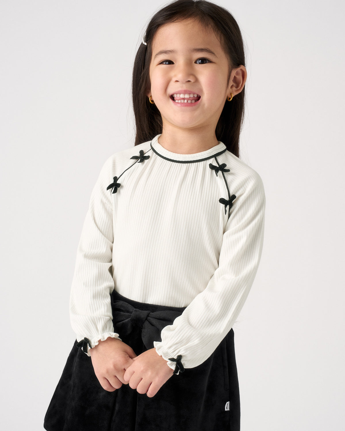 A young girl smiling, wearing a cream ribbed long-sleeve top with black bow details on the shoulders and cuffs, paired with a black skirt, against a plain light gray background.