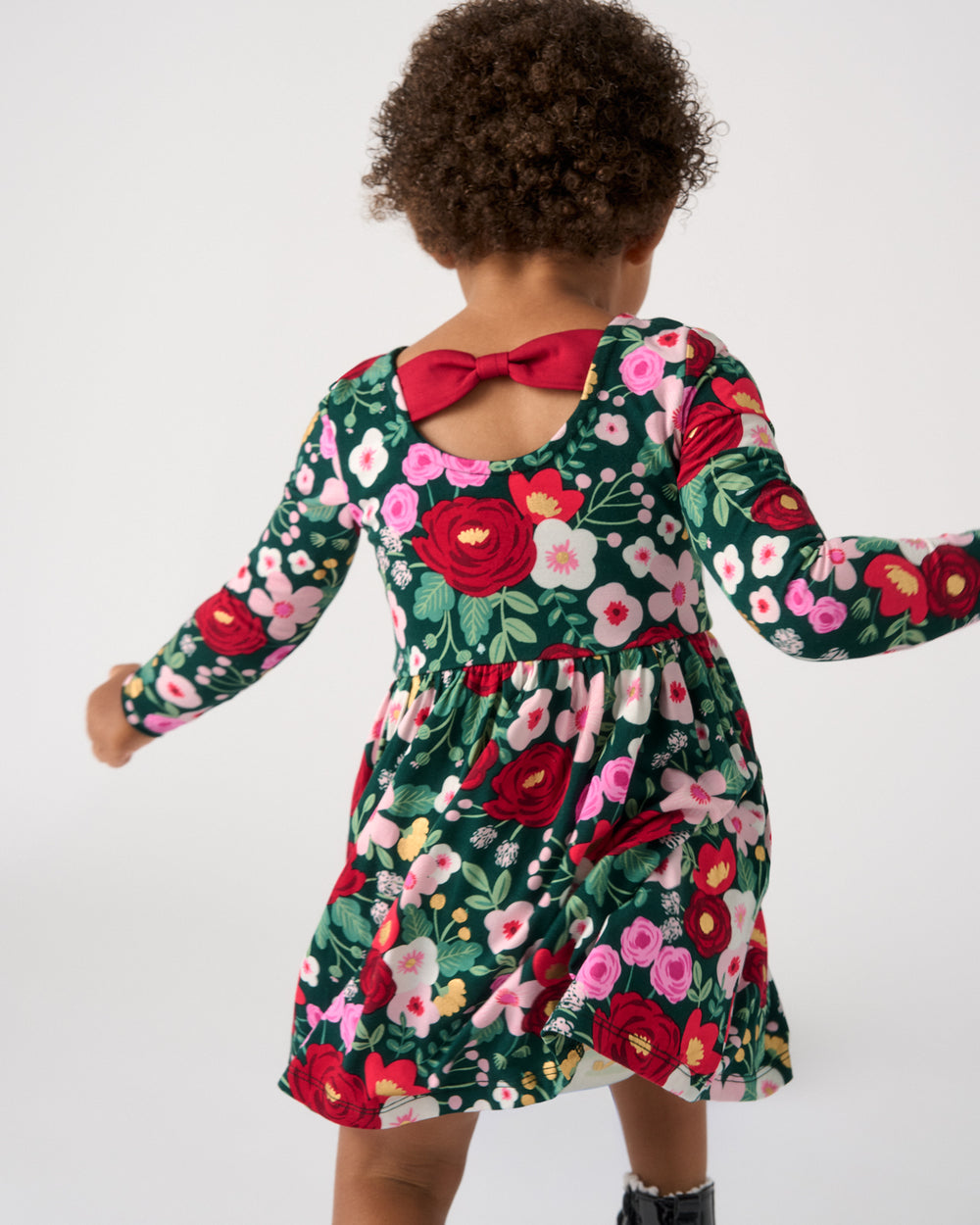 A toddler viewed from behind wearing a long-sleeve floral dress with a red bow at the back neckline, set against a plain light gray studio background.