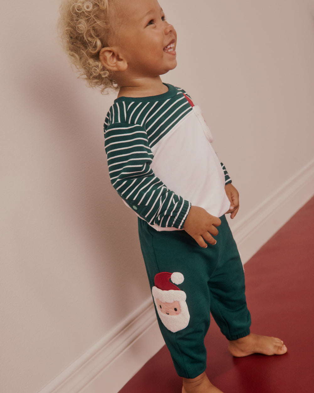 A toddler standing barefoot in a hallway, wearing a holiday-themed long-sleeve top and pants set. The top has dark green and white horizontal stripes on the sleeves and a white body panel, while the pants are dark green with a Santa Claus face applique near the lower leg.