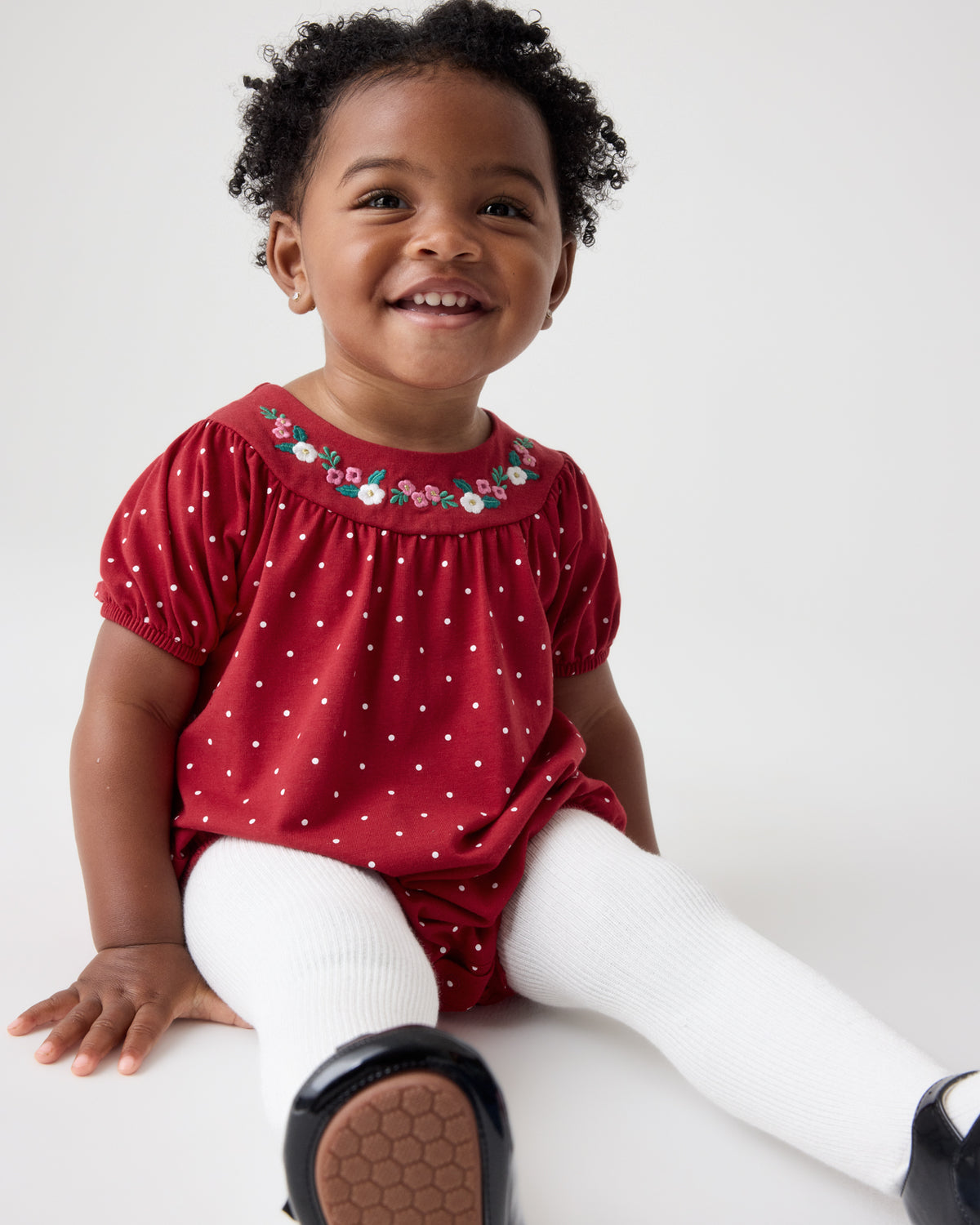 A smiling toddler girl sitting on a white floor wearing a red short-sleeve romper with white polka dots and a floral embroidered neckline, paired with white tights and black shoes.