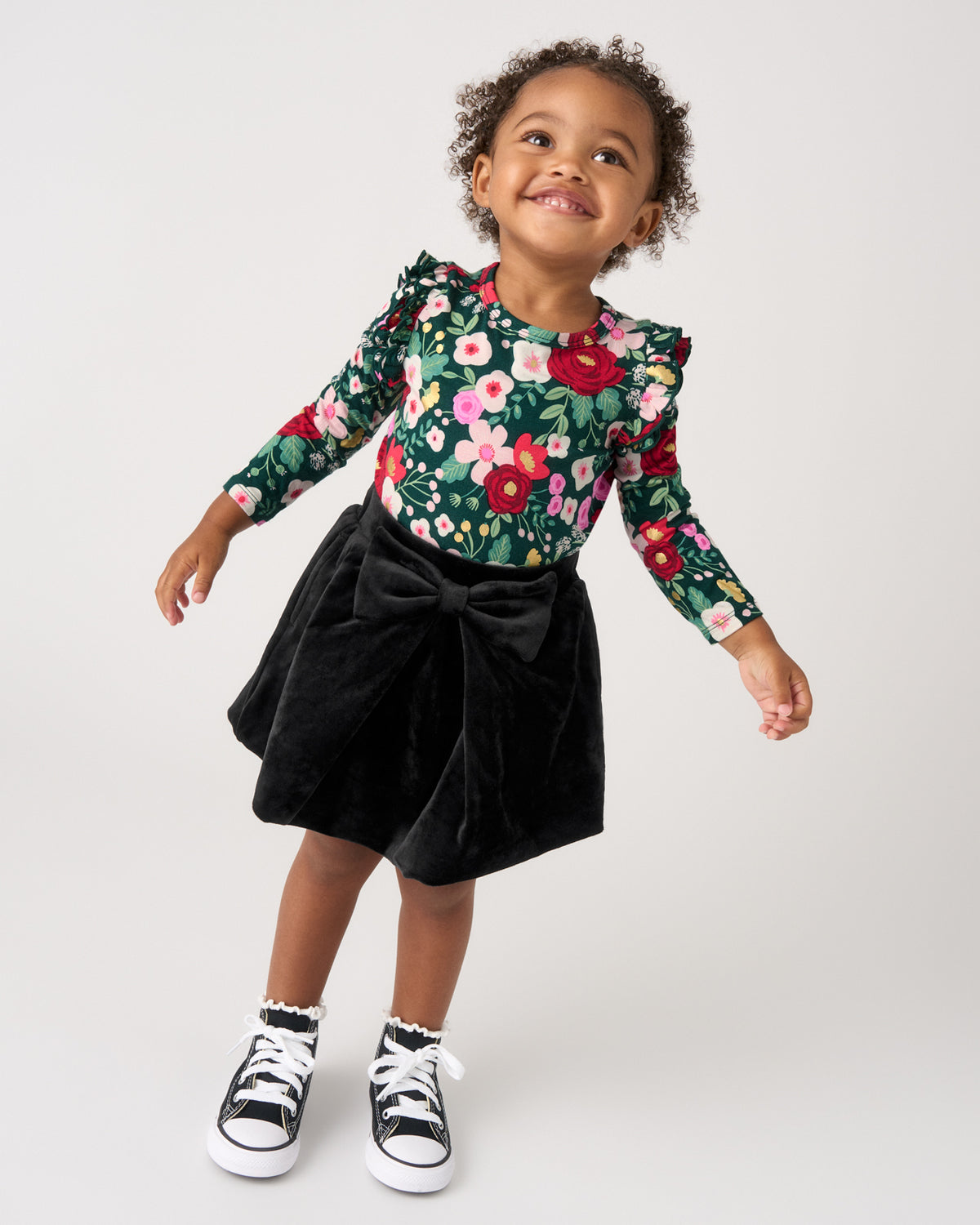 A toddler girl posing in a floral long-sleeve top with puffed shoulders and a black velvet skirt with a large bow, paired with black and white sneakers, against a light gray studio background.