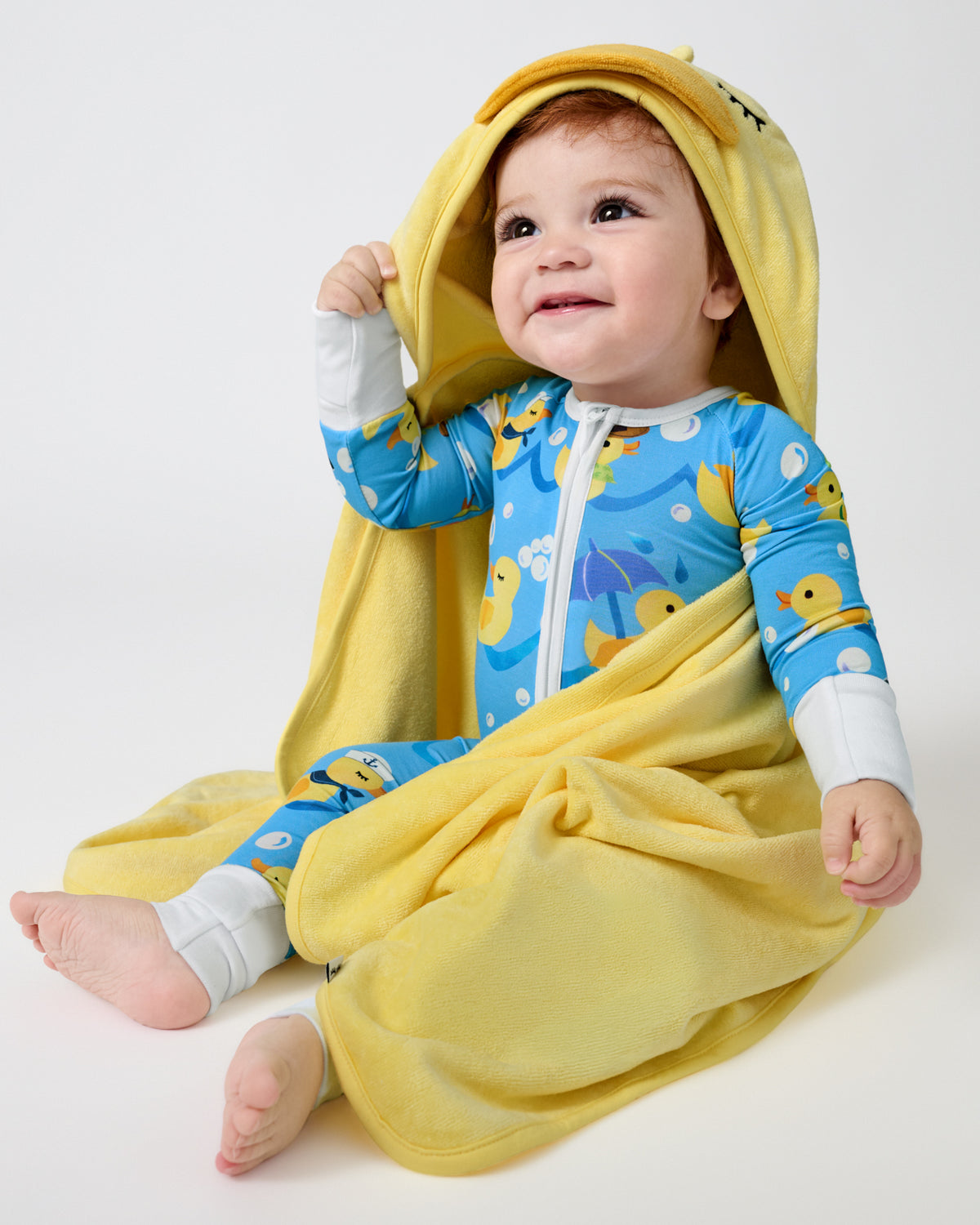 A baby wearing a blue duck-print zip-up hooded onesie and wrapped in a yellow hooded towel, sitting against a plain light gray studio background.
