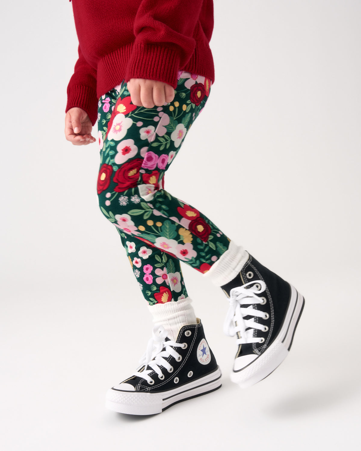 A child wearing green floral leggings, a red sweater, white socks, and black high-top sneakers, shown from the waist down against a light gray studio background.