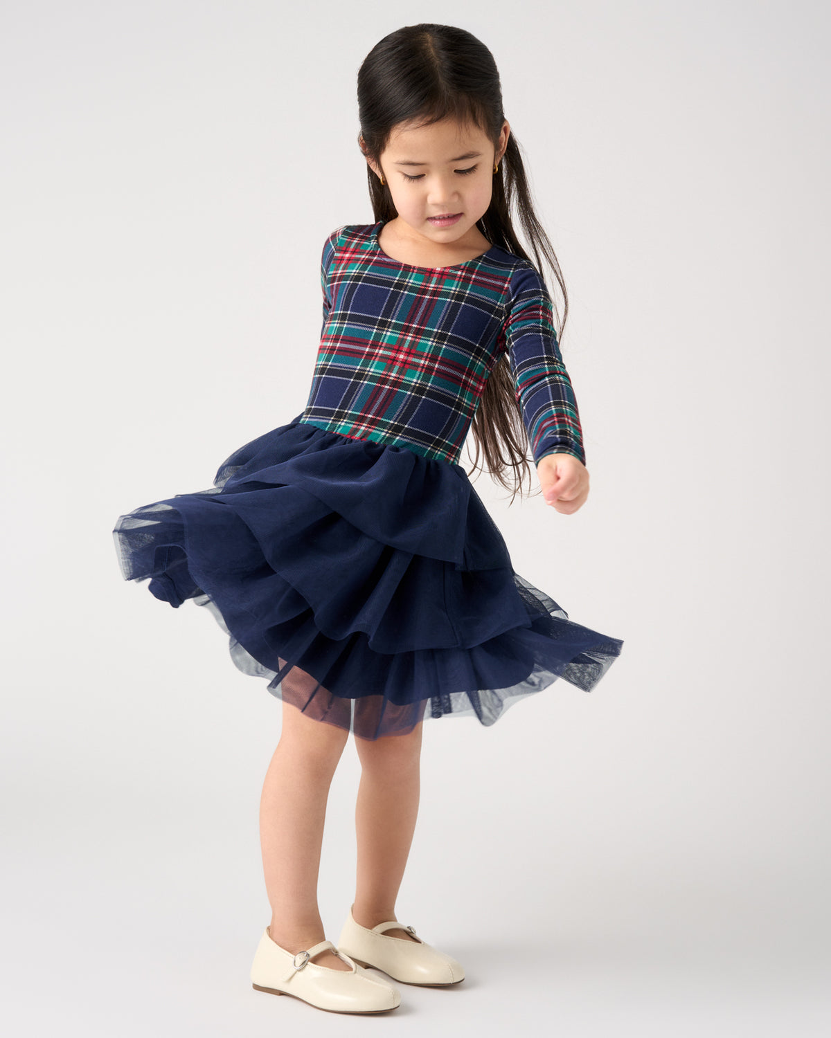 A young girl with long dark hair is wearing a navy blue dress with a green, red, and blue plaid bodice and a layered tulle skirt, paired with cream-colored shoes, against a light gray studio background.