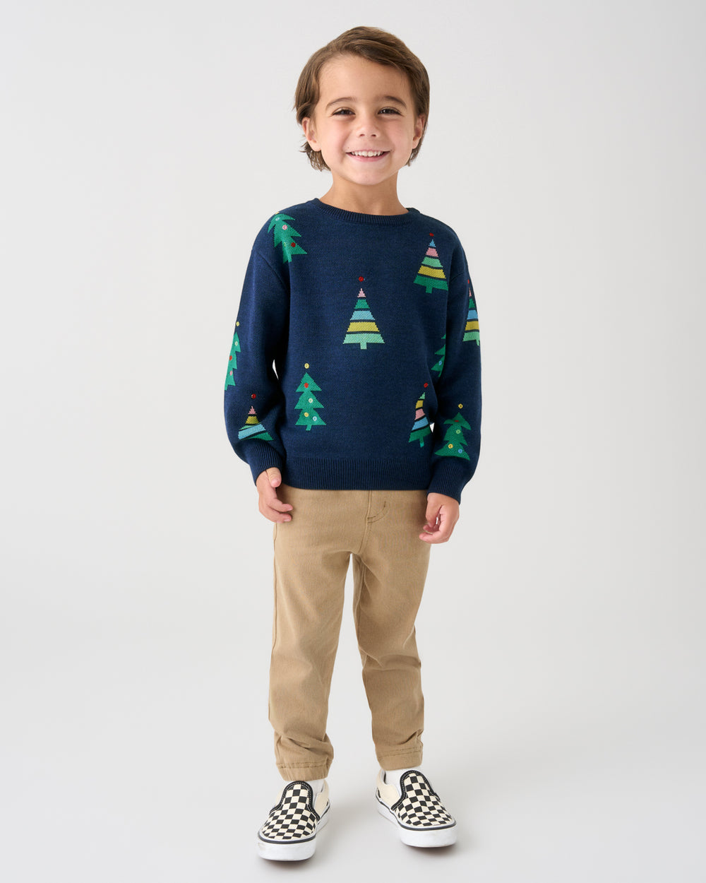 A young boy stands against a light gray studio background wearing a navy crewneck sweater with a green and yellow Christmas tree pattern, khaki pants, and black-and-white checkered slip-on shoes.