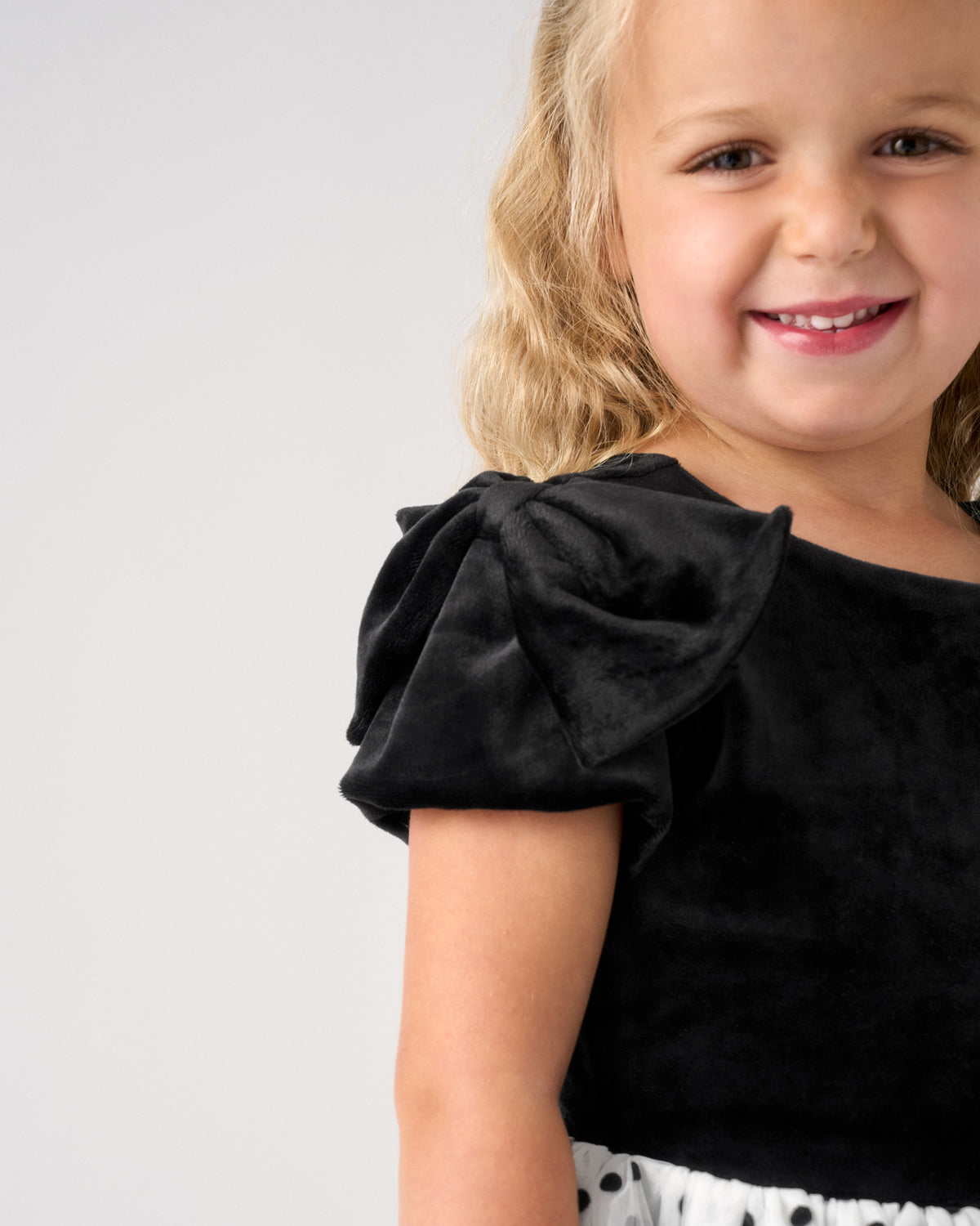 A young girl with blonde hair smiling, wearing a black velvet puff-sleeve top and a white skirt with black polka dots, photographed against a light gray studio background.
