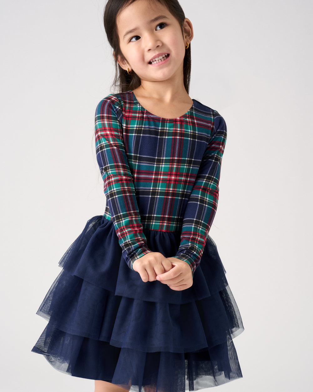 A young girl wearing a long-sleeve navy blue dress with a red, green, and blue plaid bodice and a layered navy blue tulle skirt, standing against a light gray studio background.