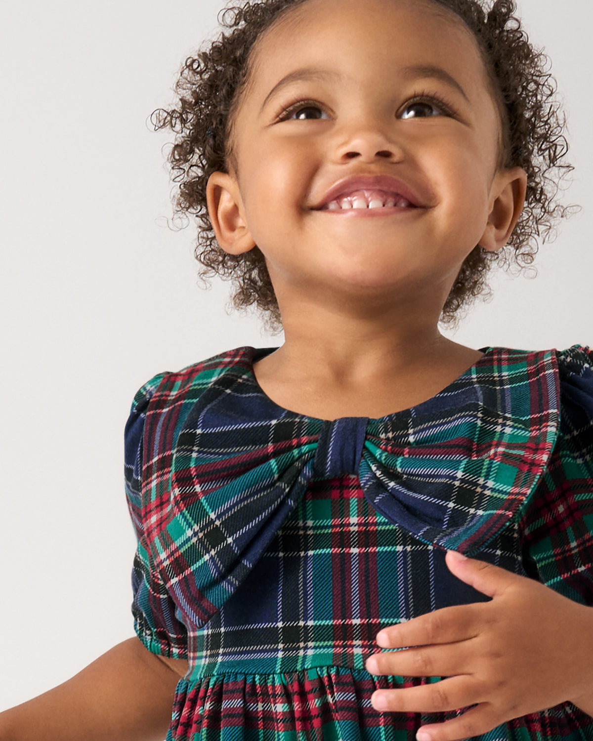 A smiling young child with curly hair wearing a navy plaid dress with a large bow at the neckline, photographed against a light gray studio background.