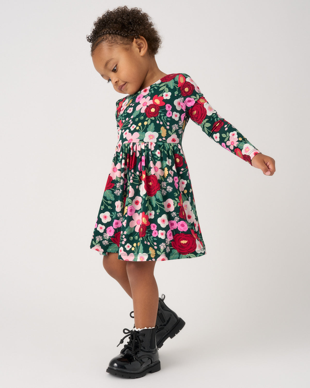 A toddler girl wearing a long-sleeve green dress with a vibrant red, pink, and white floral print, paired with black lace-up boots, posing against a light gray studio background.