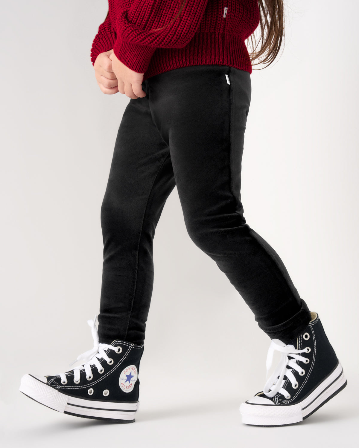 A child wearing black slim-fit pants, a red knit sweater, and black high-top sneakers with white laces and soles, shown from the torso down against a light gray studio background.