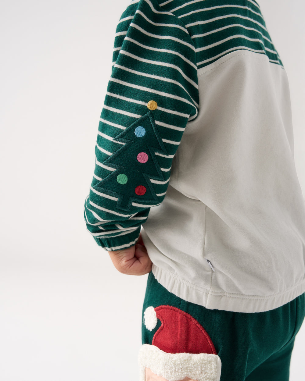 A close-up of a child wearing a dark green and white striped long-sleeve top with a felt Christmas tree applique on the sleeve, paired with dark green pants featuring a red and white Santa hat patch.