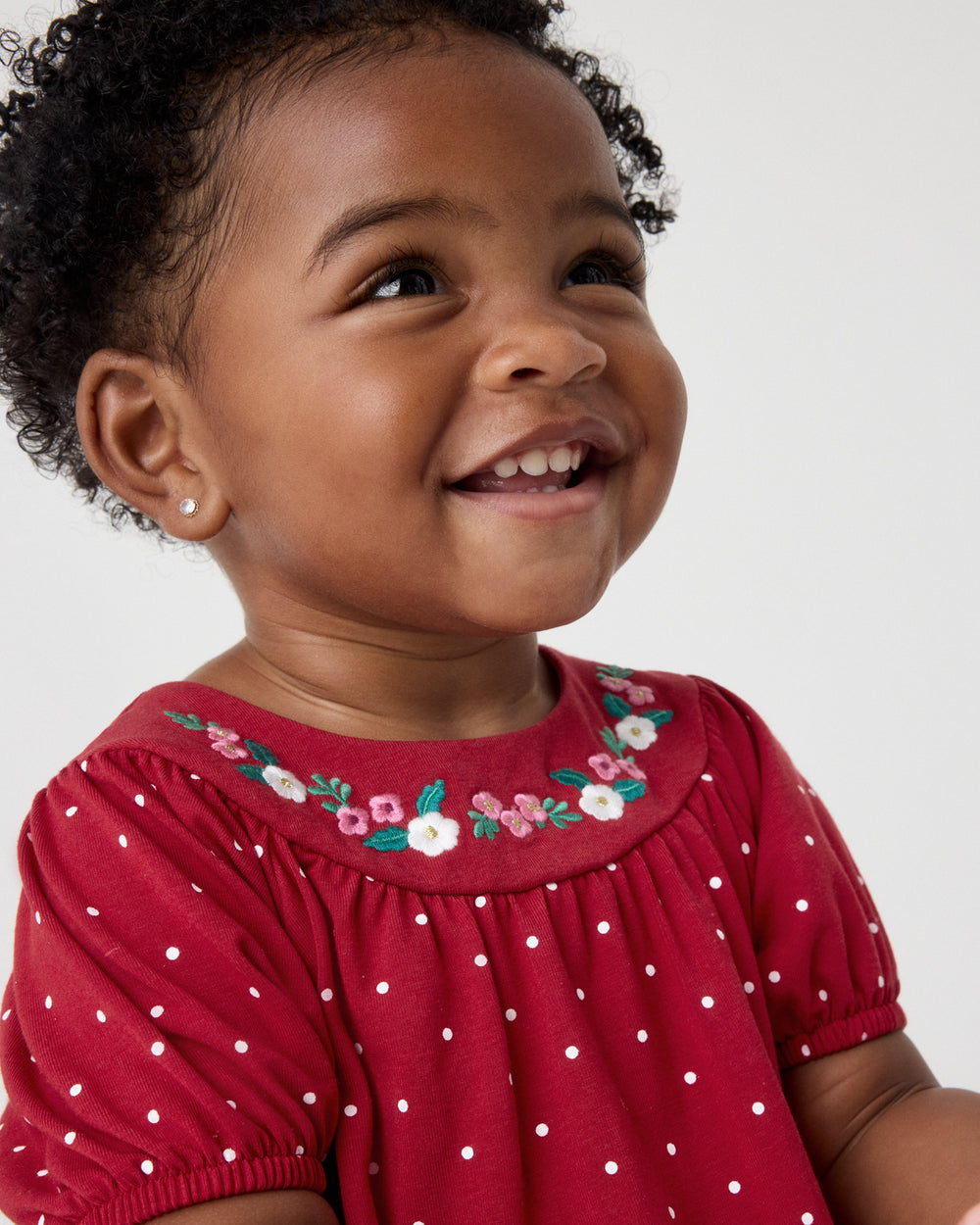 A close-up of a smiling toddler wearing a red short-sleeve top with white polka dots and a floral embroidered collar, set against a plain light gray background.