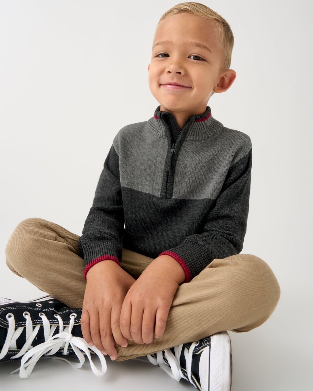 A young boy sitting cross-legged wearing a gray and charcoal quarter-zip sweater with red ribbed trim, khaki pants, and black-and-white sneakers against a plain light gray background.