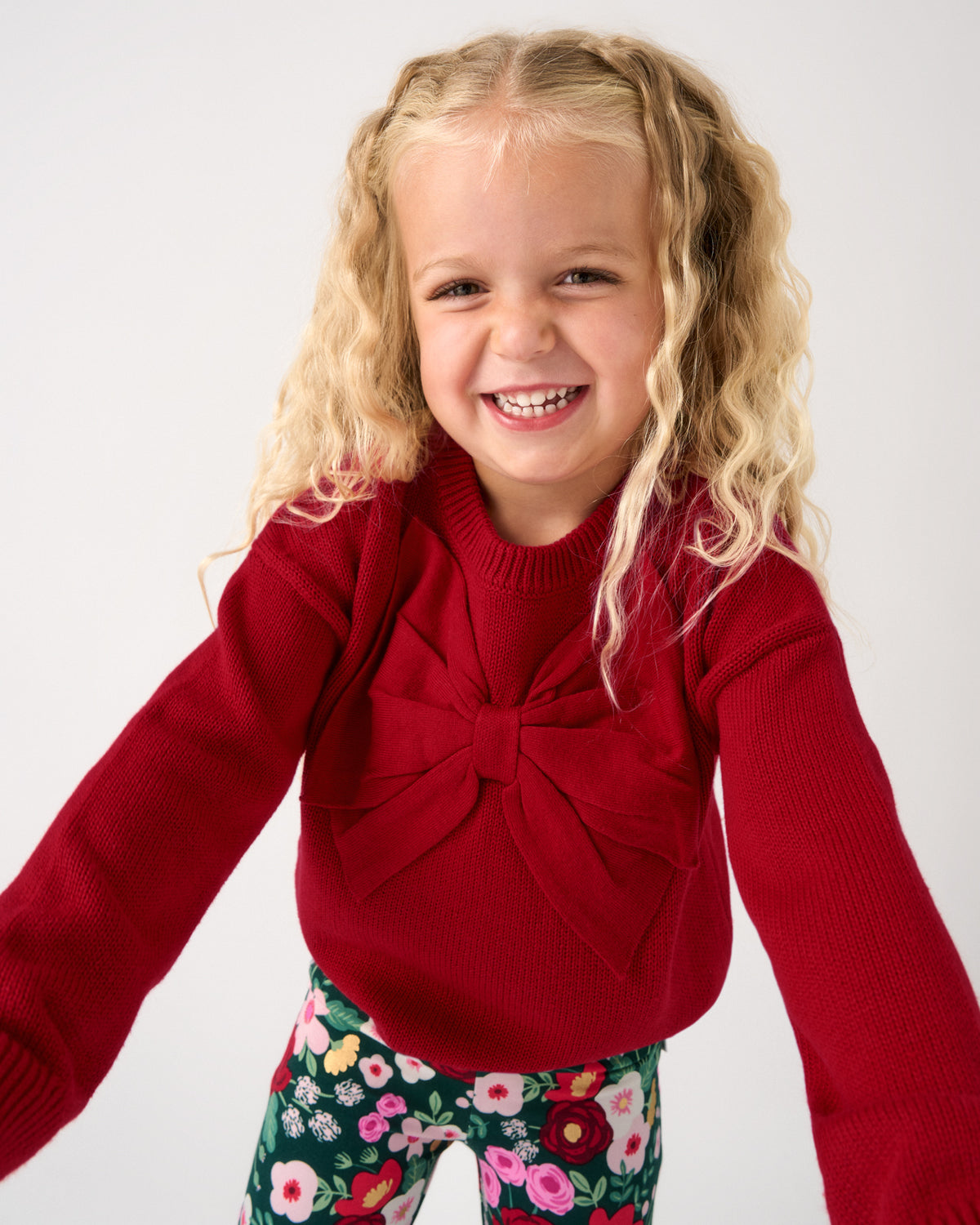 A young girl with blonde curly hair smiling, wearing a red knit sweater with a large front bow and green floral leggings, against a plain light gray background.