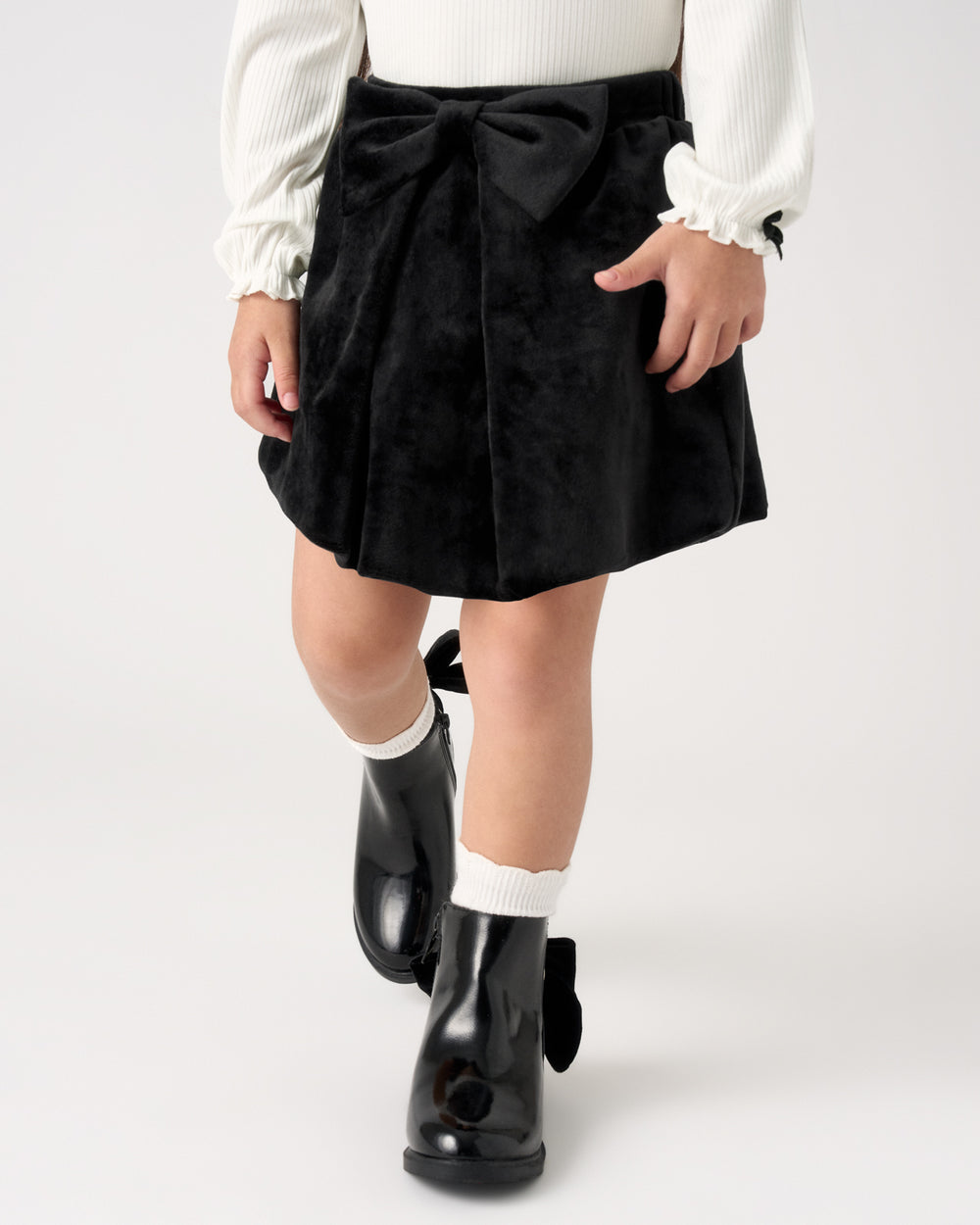 A child wearing a black velvet pleated skirt with a large bow at the waist, a white long-sleeve top, white socks, and black rain boots, shown from the waist down against a light gray studio background.